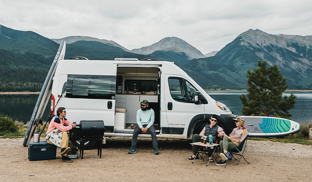 Four people sitting outside of a van with mountains in the background